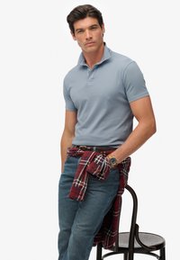 Jasnoniebieska koszulka polo, krótki rękaw, noszona z ciemnoniebieskimi dżinsami. Czerwona koszula w kratę związana w talii, złoty zegarek na nadgarstku, siedząca na krześle.