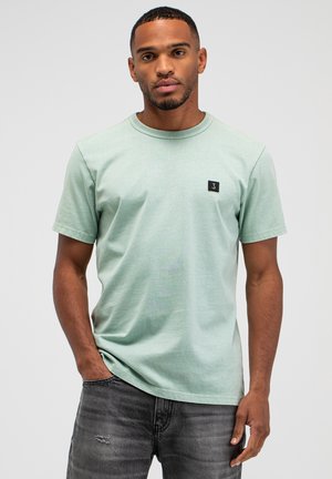 Jonge man die een lichtgroen T-shirt met korte mouwen en ronde hals draagt, met een klein zwart logo op de borst, gecombineerd met donkergrijze versleten spijkerbroeken.