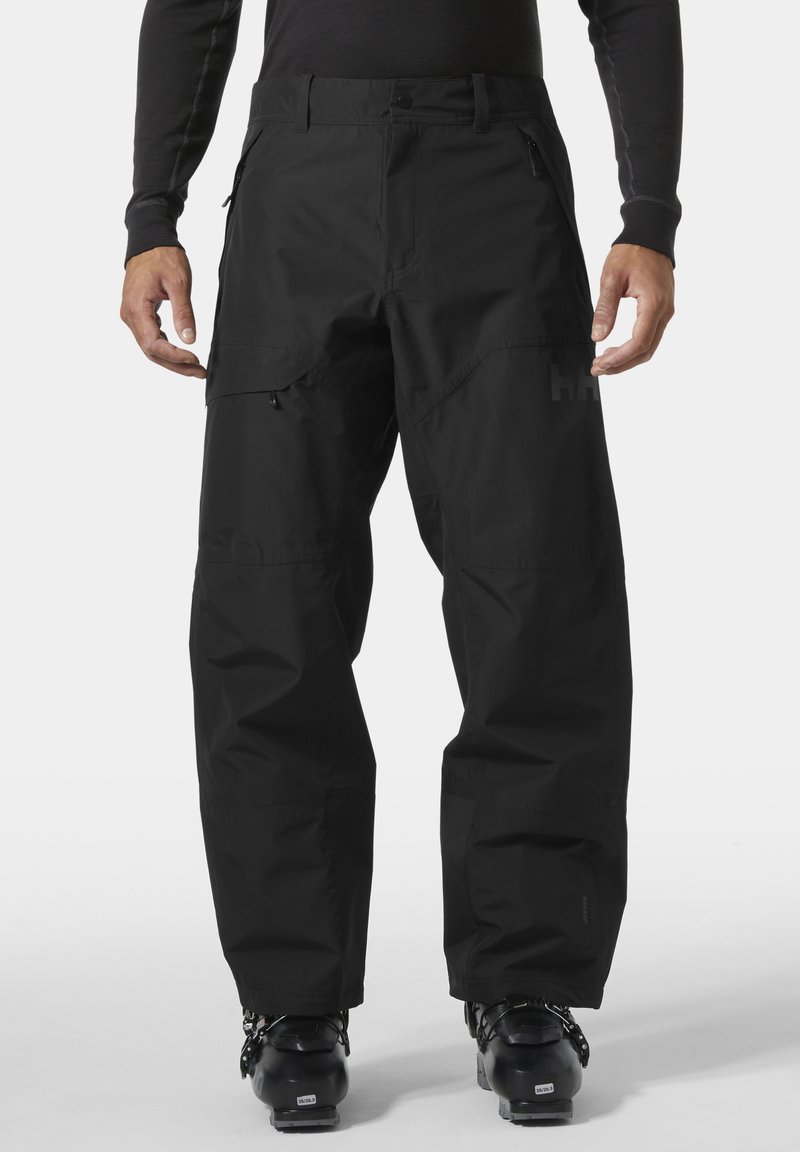 Uomo che indossa pantaloni da neve impermeabili neri e scarponi da sci neri, in piedi davanti a uno sfondo bianco uniforme.