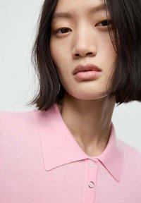 Helles rosa Poloshirt mit weicher Textur. Verfügt über zwei kreisförmige silberne Knöpfe am Kragen. Der Hintergrund ist neutral.