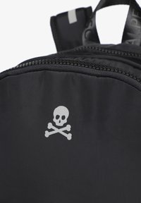 Mochila negra de nailon con un prominente gráfico de calavera y huesos cruzados en blanco, textura suave y detalles de cremallera en contraste.
