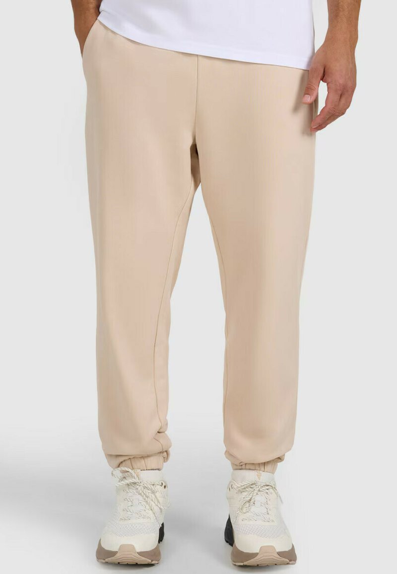 Osoba ubrana w beżowe spodnie dresowe z elastycznymi mankietami i białe trampki, stojąca na tle jasnoszarego tła.