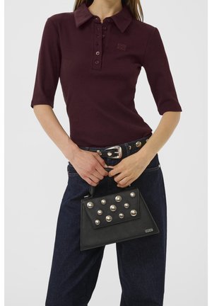 Vrouw die een donkerpaars poloshirt en donkere spijkerbroek draagt, houdt een zwarte handtas met grote ronde zilveren studs vast.