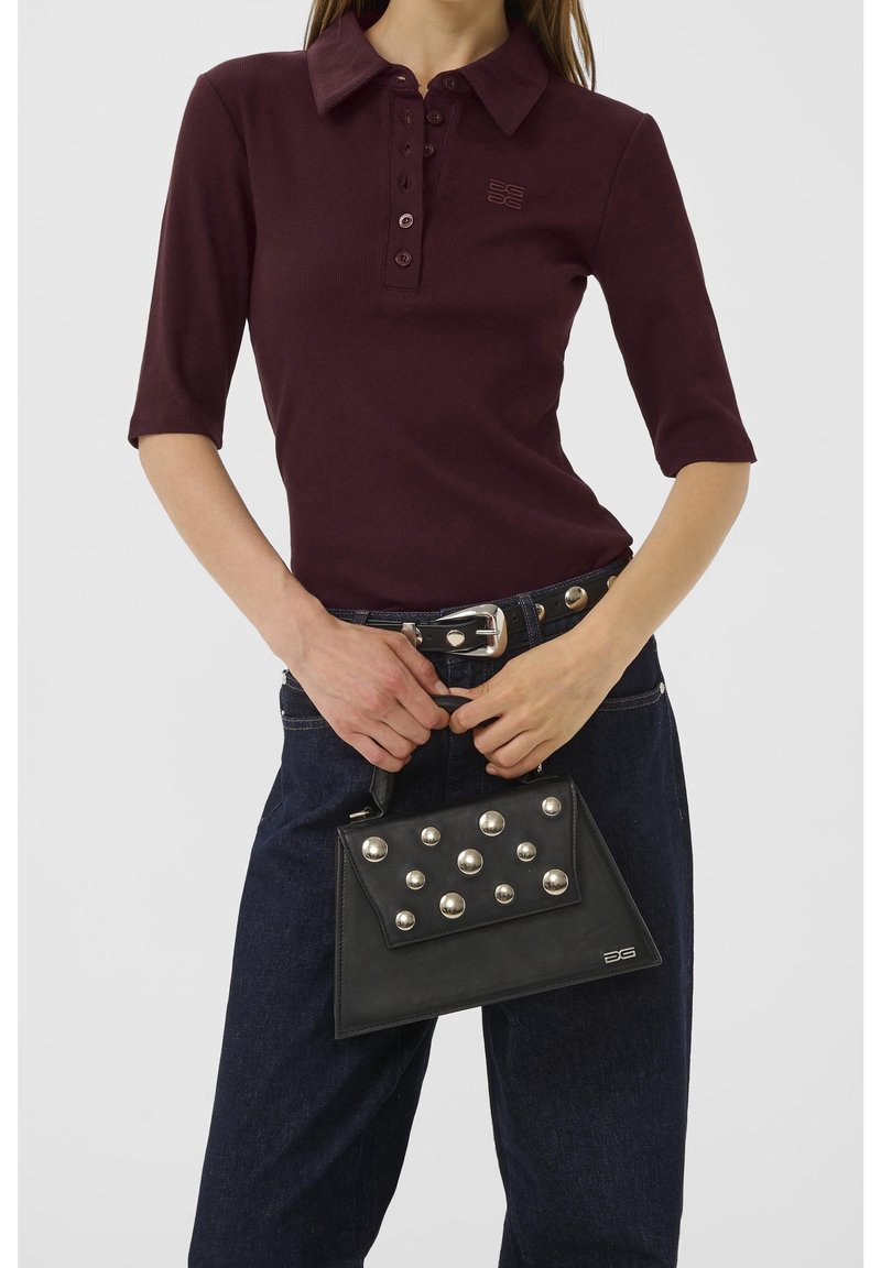 Femme portant un polo violet foncé et un jean foncé, tenant un sac à main noir avec de gros clous ronds argentés.