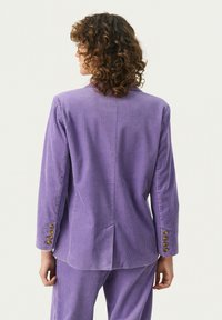 Violetti corduroy-takki, jossa on pystysuorat raidat, yksinkertainen halkio takana ja koristeelliset kultanapit hihoissa.