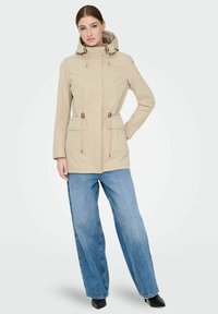 Veste beige à capuche, avec deux poches avant, taille cintrée et cordon de serrage. Associée à un jean bleu à jambes larges et des chaussures noires.