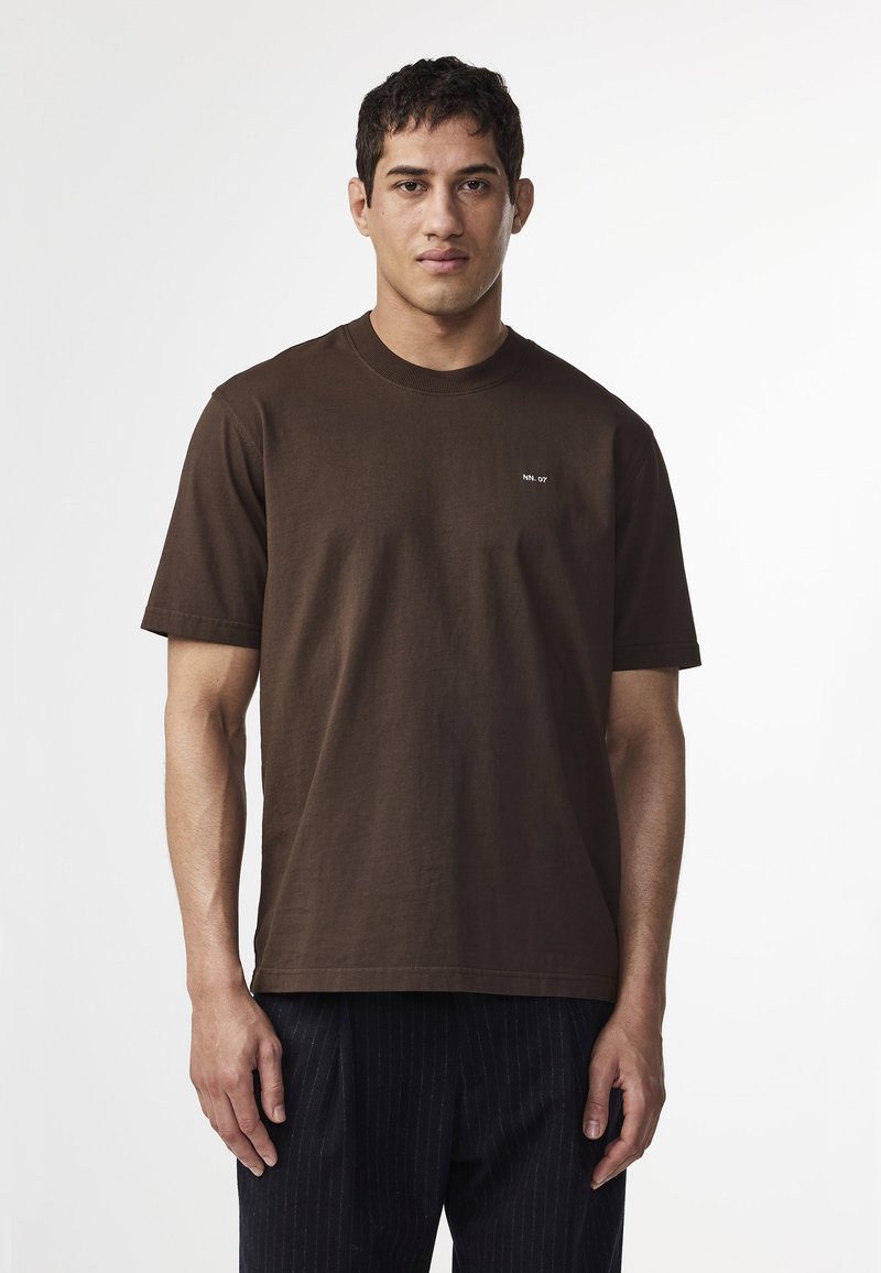 Man i en enkel mörkbrun kortärmad t-shirt med liten vit text på bröstet, tillsammans med mörka ränderbyxor, mot vit bakgrund.