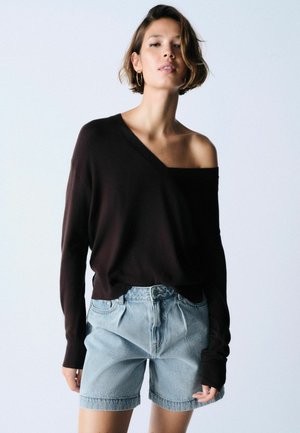 Femeie cu păr scurt care poartă un pulover negru cu umerii goi și pantaloni scurți din denim bleu deschis cu talie înaltă pe un fundal simplu.