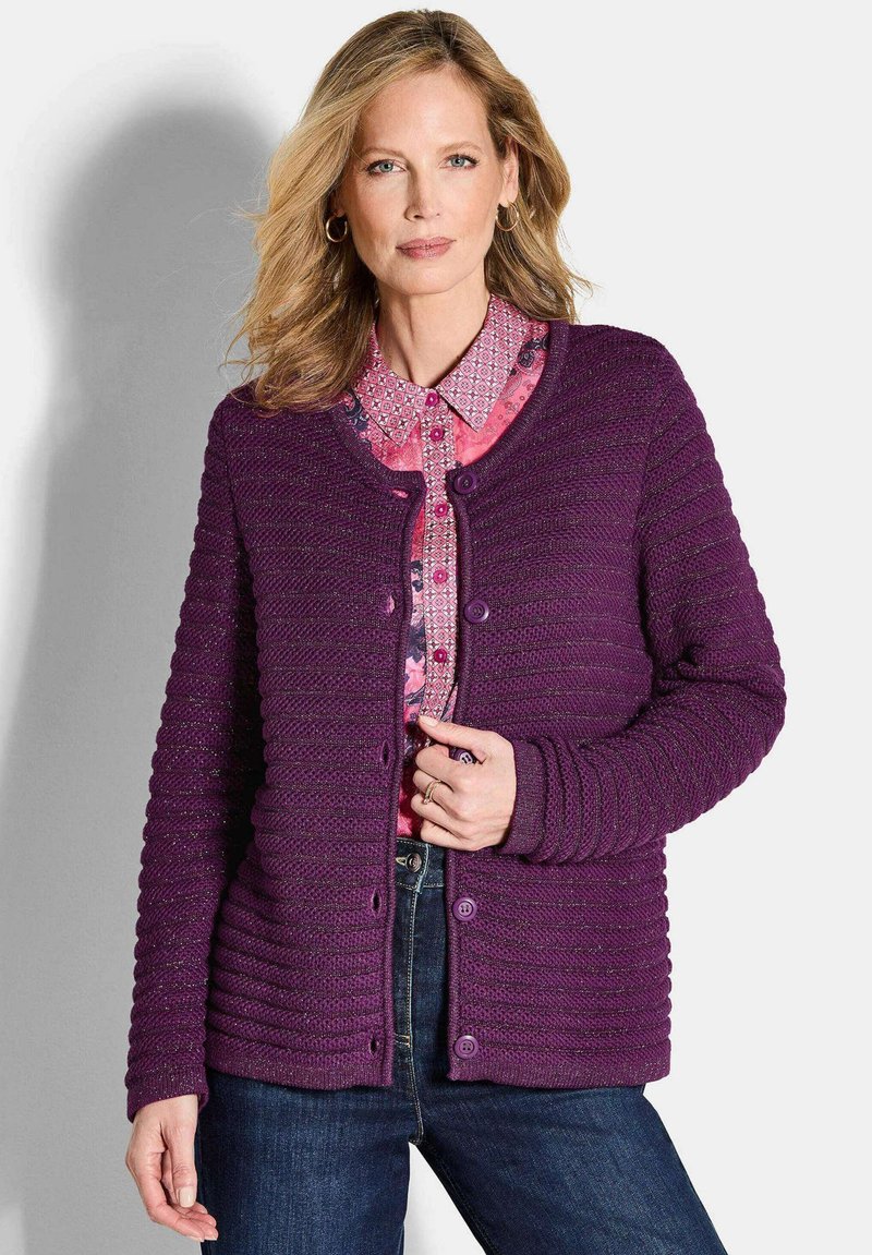 Frau trägt eine lila, geknöpfte Strickjacke über einem rosa gemusterten Shirt und dunklen blauen Jeans, steht vor einem hellen Hintergrund.