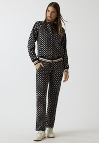 Zwarte trainingspak met een herhalend geometrisch patroon in crème. Voorzien van een nauwsluitend jack met rits en geribbelde manchetten, gecombineerd met bijpassende broek.