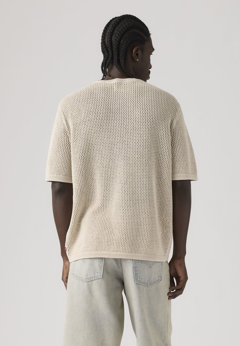 Homme aux cheveux tressés portant un pull court beige en maille ajourée et un jean délavé clair, tourné de dos sur un fond uni.