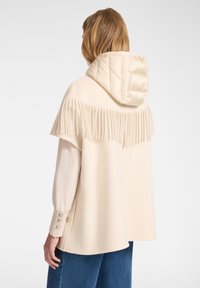 Cappotto beige con cappuccio voluminoso, dotato di dettagli frangiati sulle spalle e bottoni decorativi sulle maniche. Tessuto morbido e testurizzato.