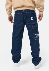 Mörkblå denim cargo-byxor med bakfickor, med vita logotyper och textdetaljer. Löst skuren för komfort, med synliga sömnadsdetaljer.