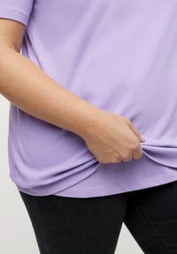 Svetle fialový top s krátkými rukávy a hladkým povrchem, stažený v pase, v kombinaci s černými upnutými kalhotami. Detailní záběr na ruku, která tahá za látku.