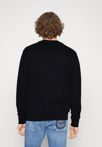 Svart sweatshirt med ribbad mudd och nederkant, enkel design, bärs med ljusblå jeans som har en cirkulär logotyp-patch på bakfickan.