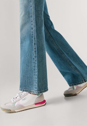 Persona con vaqueros azules y zapatillas blancas con detalles en rosa y verde, caminando sobre una superficie lisa de color gris claro.