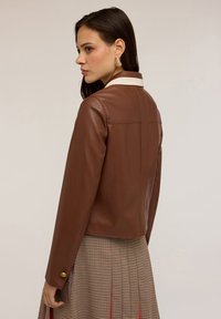 Giovane donna con giacca di pelle marrone con colletto bianco, che indossa orecchini d'oro e una gonna a quadri con pieghe rosse, rivolta di lato.
