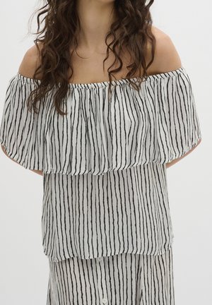 Vrouw draagt een witte off-shoulder blouse met verticale zwarte strepen en een bijpassende gestreepte rok, met golvend donker haar.