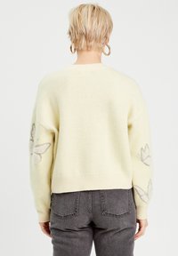 Lysegul strikket sweater med lange ærmer, har broderede sommerfugledesign på begge ærmer og en ribbet kant.