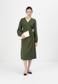 Woman in a long-sleeve olive green wrap dress holding a cream clutch, wearing black-and-beige slingback flats, standing on a white background.