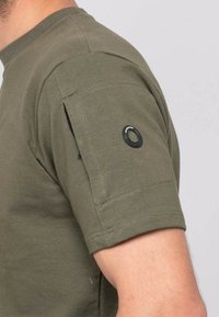 T-shirt à manches courtes vert olive en tissu doux. Doté d'une poche avec une fermeture éclair sur la manche et d'un détail logo noir circulaire.