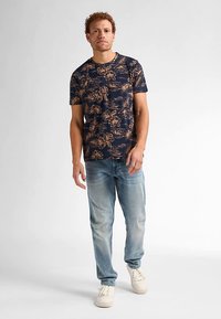 Camiseta de manga corta azul marino con un patrón floral en tonos beige, combinada con vaqueros azul claro y zapatillas blancas, posando frente a un fondo liso.