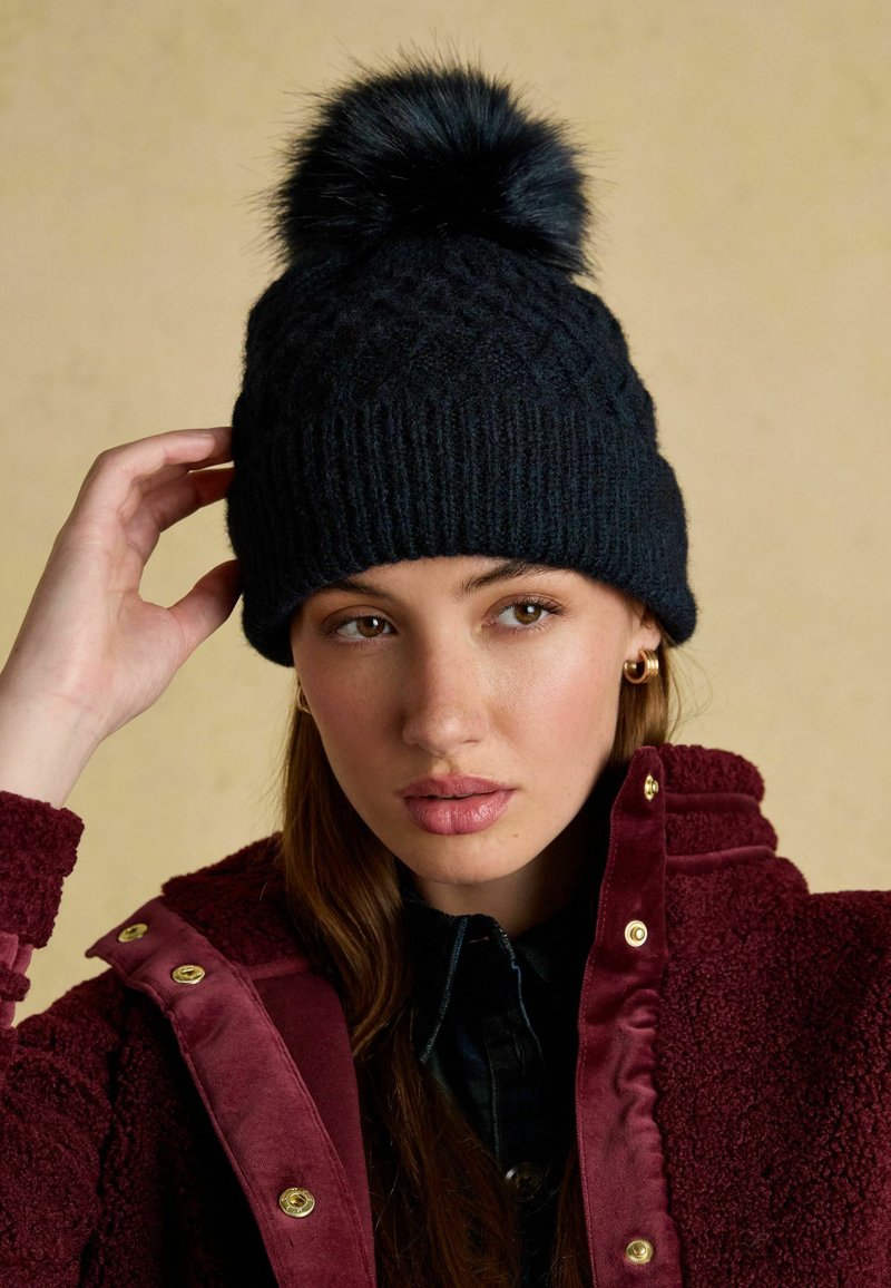 Dark navy knitted beanie with a fluffy black pom-pom, featuring a textured pattern, worn by a person against a neutral background.