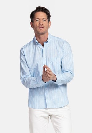 Man met kort bruin haar, gekleed in een lichtblauw overhemd met verticale strepen en witte broek, met de handen gevouwen voor zich.