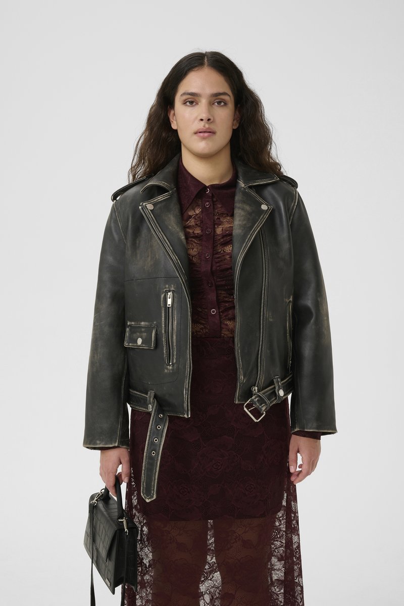 Μαύρο δερμάτινο μπουφάν μοτοσικλέτας με φθαρμένη επιφάνεια, ασημένια μεταλλικά στοιχεία και πολλές τσέπες. Φοριέται πάνω από μια μπορντό δαντελένια φορεσιά, κρατημένο με μια μαύρη τσάντα.