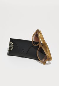 Ochelari de soare din plastic transparent maro cu lentile întunecate, plasați parțial într-un toc texturat negru cu un logo.