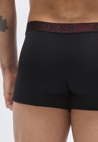 Černé trenýrky z měkké látky, s širokým vínovým pasem s logem "GUESS", hladkou texturou a přiléhavým střihem.