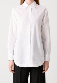 Persona che indossa una camicia bianca semplice con colletto e maniche lunghe, abbinata a pantaloni neri su uno sfondo neutro.