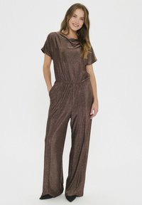 Bronz színű fémes overál rövid ujjakkal, aszimmetrikus dekoltázssal, rugalmas derékkal, széles lábakkal és oldalsó zsebekkel. Simább, fényes textúrájú.