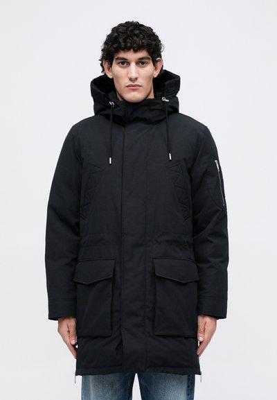 Vyras, vilkintis juodą paminkštintą parka su kapišonu, priekinėmis kišenėmis ir užtrauktukais ant rankovių, stovi prieš baltą fono.