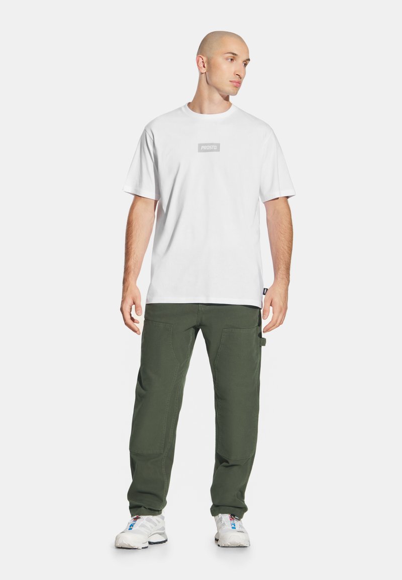 Mężczyzna w białej koszulce "PROSTO.", oliwkowych spodniach cargo i białych sneakersach, stojący na tle czystego białego tła.