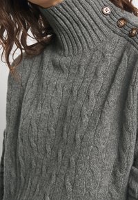 Pull en maille grise avec col montant côtelé, orné de trois boutons marron sur une épaule. Texture douce et design épais.
