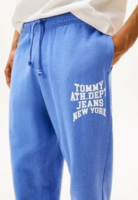 Blaue Jogginghose aus weichem Material, mit einem Kordelzug im Bund und einem weißen Aufdruck mit dem Design "TOMMY ATH.DPT JEANS NEW YORK" an der Seite.