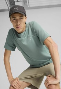 Heledad roheline, tekstuuriga lühikeste varrukatega spordisärk PUMA logoga; koos beeži lühikesed püksid. Must müts PUMA logoga ees.
