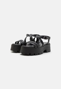 Black platform sandals feature multiple broad straps, an adjustable ankle buckle, and a textured, chunky sole for enhanced grip.
