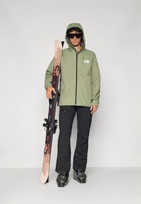 Zelena vodoodporna jakna z kapuco, črne smučarske hlače in škornji. Drži smučko z bež osnovo in rdečimi poudarki. Na glavi ima temna sončna očala.