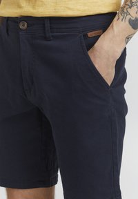 Marineblaue Shorts aus Baumwolle, mit Seitentaschen, einem Knopfverschluss und einem braunen Lederetikett am Bund.