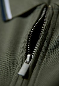 Mörkolivgrön tyg med en närbild av en silverblixtlås, som har en texturerad yta och metalltänder.