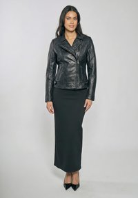 Kobieta stojąca w czarnej skórzanej kurtce, długiej czarnej spódnicy i czarnych szpilkach z ostrym noskiem na jednolitym jasnym tle.