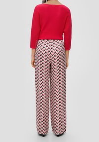 Donna che indossa una maglietta rossa a costine e pantaloni larghi a vita alta con un motivo a zigzag rosa, nero e bianco, in piedi di spalle.