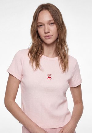 Jeune femme aux cheveux châtain clair portant un t-shirt côtelé rose pâle avec un petit motif de cerise et un texte au centre de la poitrine.