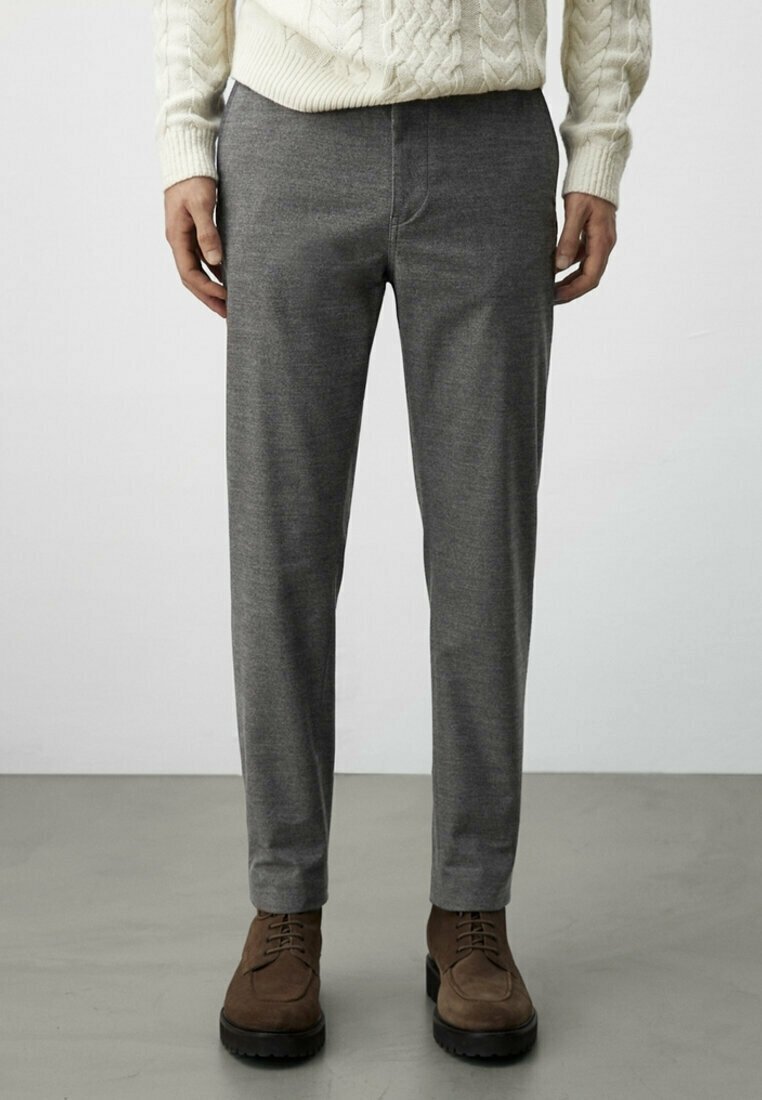 Homme portant un pantalon gris slim, un pull en maille torsadée crème et des bottes marron à lacets, debout sur un sol gris.