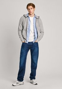 Sudadera gris con cremallera sobre una camisa de botones a rayas azul claro, camiseta blanca, jeans azules y zapatillas grises con detalles. Conjunto sencillo y casual.