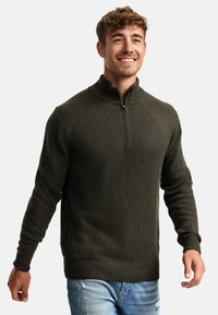 Hombre sonriente vestido con un suéter gris oscuro de cremallera hasta el cuarto y jeans rasgados de color azul claro, de pie con los brazos ligeramente extendidos ante un fondo claro.