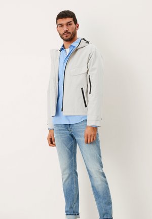 Light gray, water-resistant jacket with a hood, front zip pockets, and contrasting black zippers, worn over a blue button-up shirt and jeans.