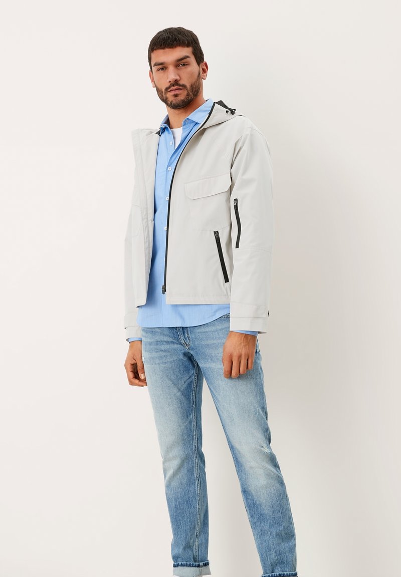 Helle graue, wasserabweisende Jacke mit Kapuze, vorderen Reißverschlusstaschen und kontrastierenden schwarzen Reißverschlüssen, getragen über einem blauen Hemd mit Knöpfen und Jeans.
